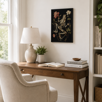A framed black-and-ivory skeleton frog artwork hangs on a white wall above a wooden desk with a lamp, open book, and books in a bright home office setting.