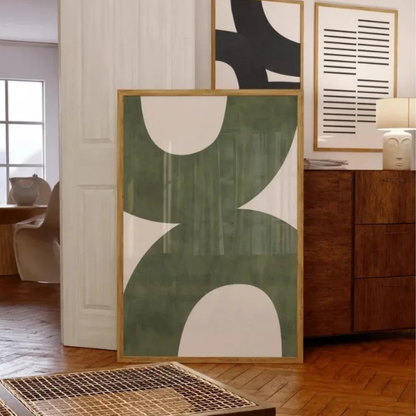 A framed abstract art print with organic shapes in sage green and cream, leaning against a white paneled wall in a modern living room setting.