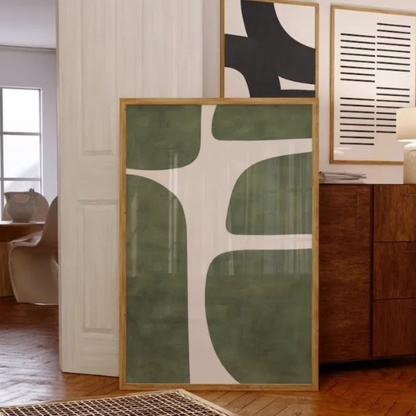 A large framed abstract art print with organic shapes in sage green and white, leaning against a white paneled wall in a modern living room setting.