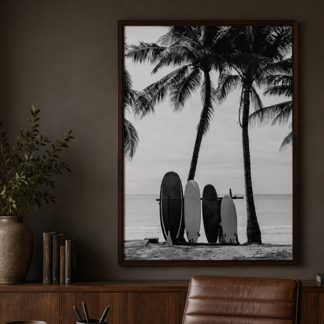 A framed black and white beach art print featuring palm trees and surfboards, hanging on a dark brown wall above a wooden console table with books and a plant.
