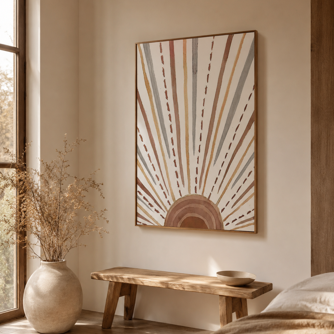 A framed wall art print featuring a sunburst design in warm beige, brown, muted blue-gray, and terracotta tones, displayed above a wooden bench in a softly lit room with a vase of dried branches nearby.