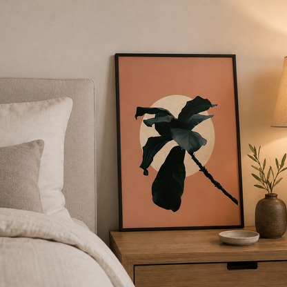 A framed botanical art print featuring a dark green leaf silhouette against a peach and cream circular background, styled on a wooden nightstand next to a bed in a warm, modern bedroom.