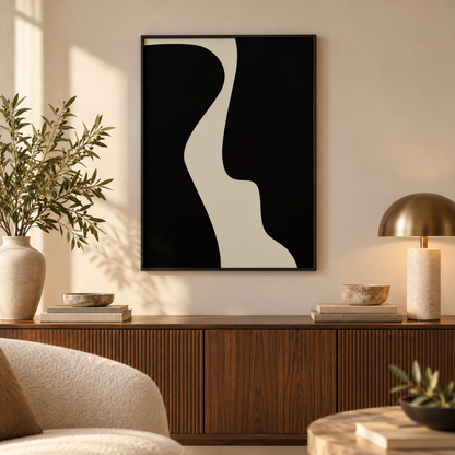A framed abstract black and off-white wall art print hangs above a wooden console in a styled living room with a lamp, bowls, books, and a plant.