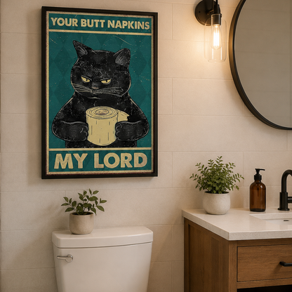 A framed humorous wall art print featuring a grumpy black cat holding a roll of toilet paper, with the text 'YOUR BUTT NAPKINS MY LORD' on a teal background, displayed in a bathroom above a toilet and next to a vanity.