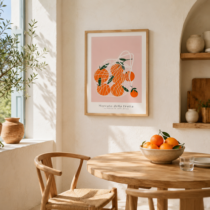 A framed art print featuring a minimalist orange illustration on a dusty pink background, hanging on a warm cream wall in a sunlit, rustic dining room with a wooden table and chairs.