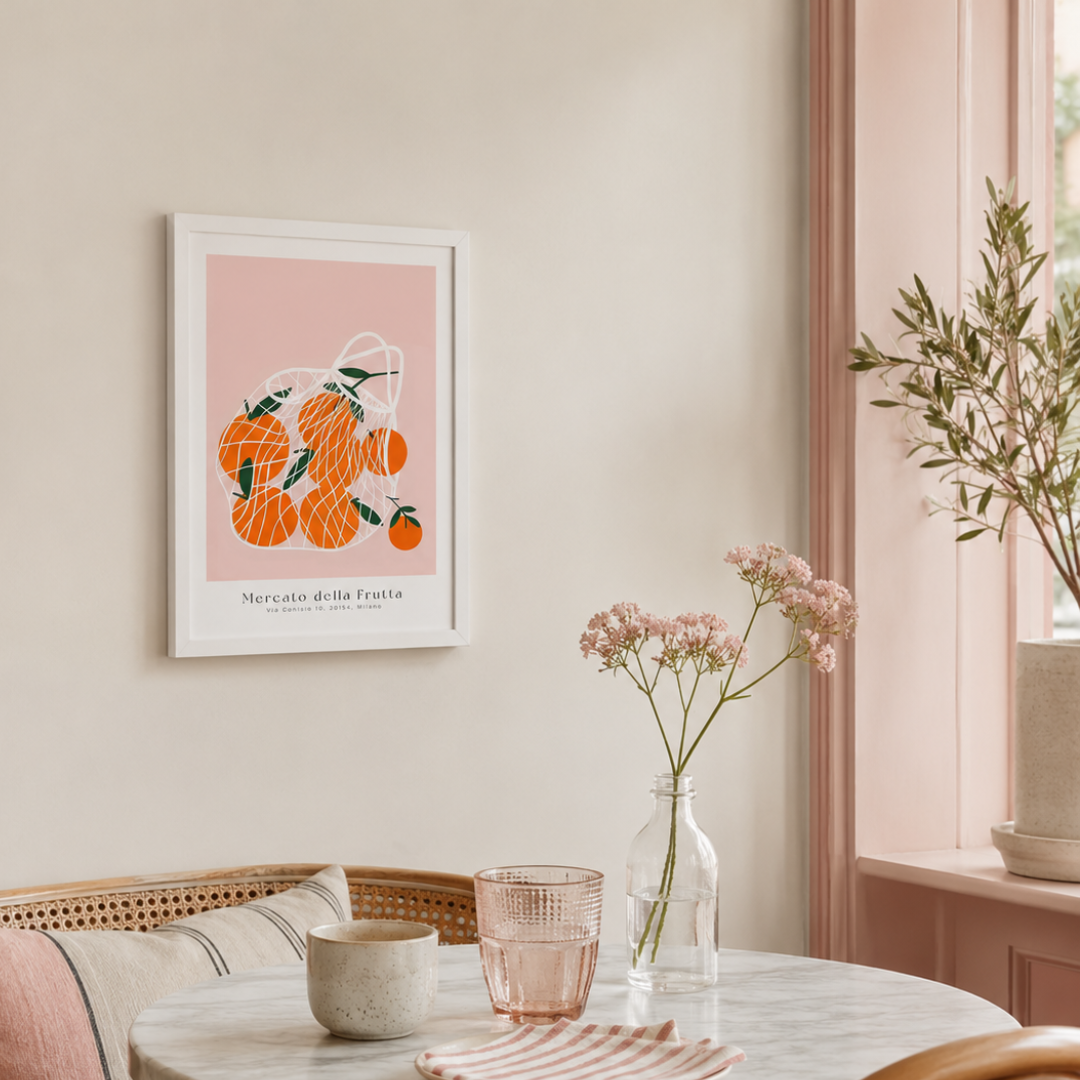 A framed art print featuring a stylized orange illustration with the Italian text 'Mercato della Frutta' hangs on a beige wall in a softly lit room. The scene includes a round marble table with a pink glass, a small bowl, and a glass vase with pink flowers, alongside a window and potted plants.