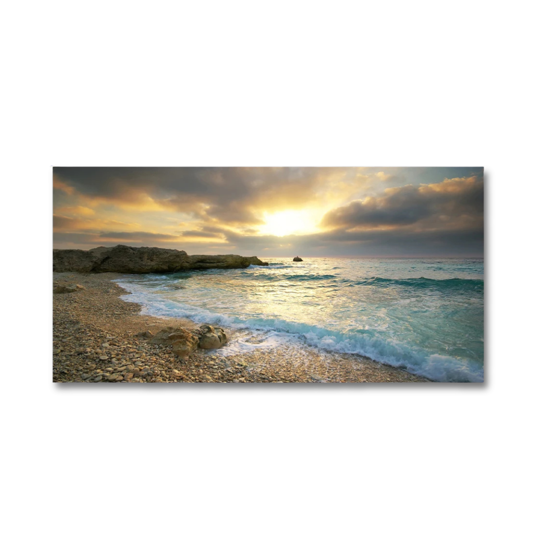 A rectangular wall art print showing a coastal beach and sea at sunset, with golden light on the water, blue-green waves, rocky shoreline, and dramatic clouds. The print is displayed against a plain white background.