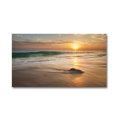A rectangular wall art print showing a beach sunset with waves, sand, and a bright sun on the horizon, displayed against a plain white background.