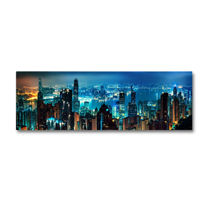 A panoramic canvas wall art print depicting a vibrant city skyline at night with illuminated skyscrapers and water, shown hanging on a plain white wall.