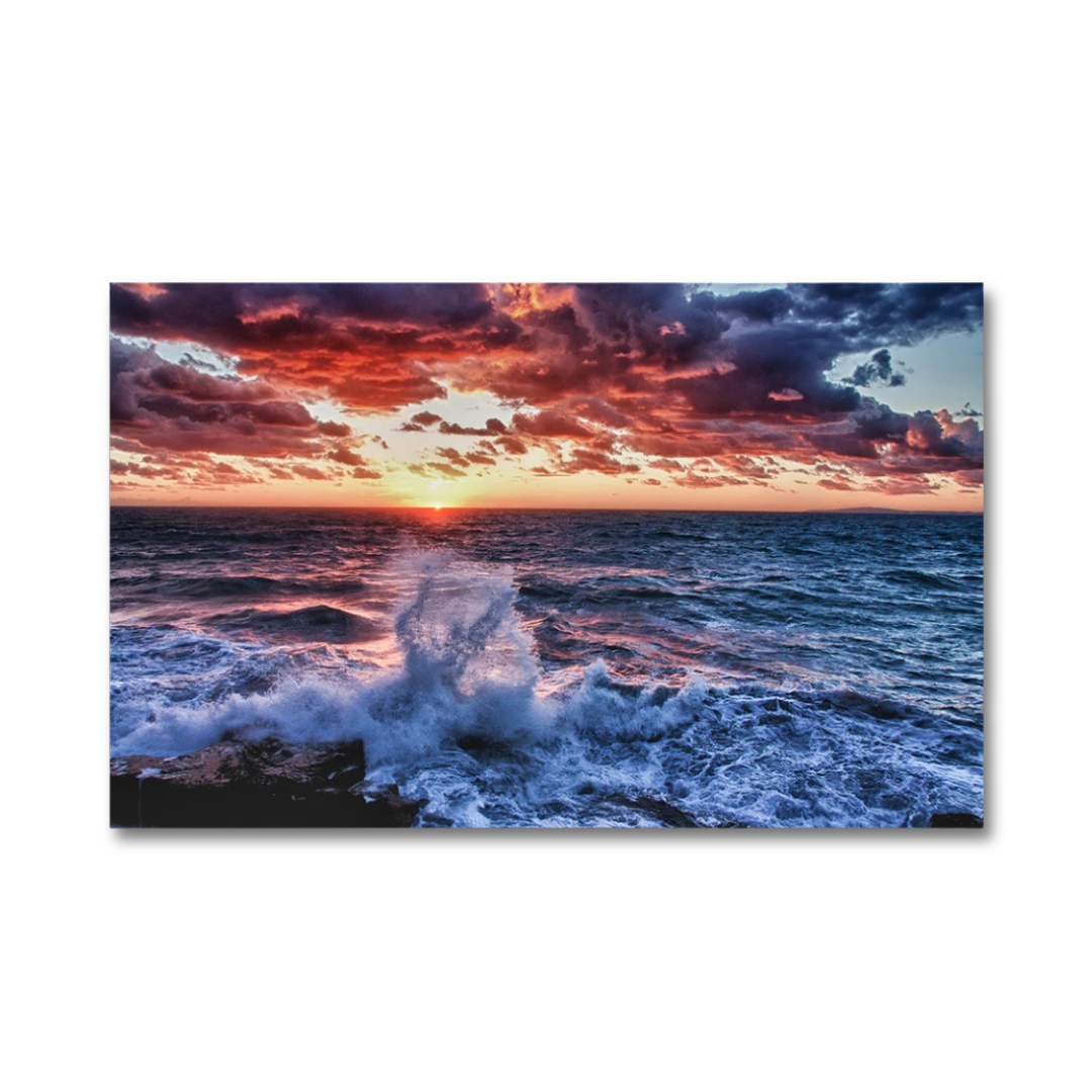 A rectangular wall art print or canvas showing a dramatic ocean sunset with dark blue water, white surf, and a fiery sky in red, orange, and purple tones, displayed against a light gray background.