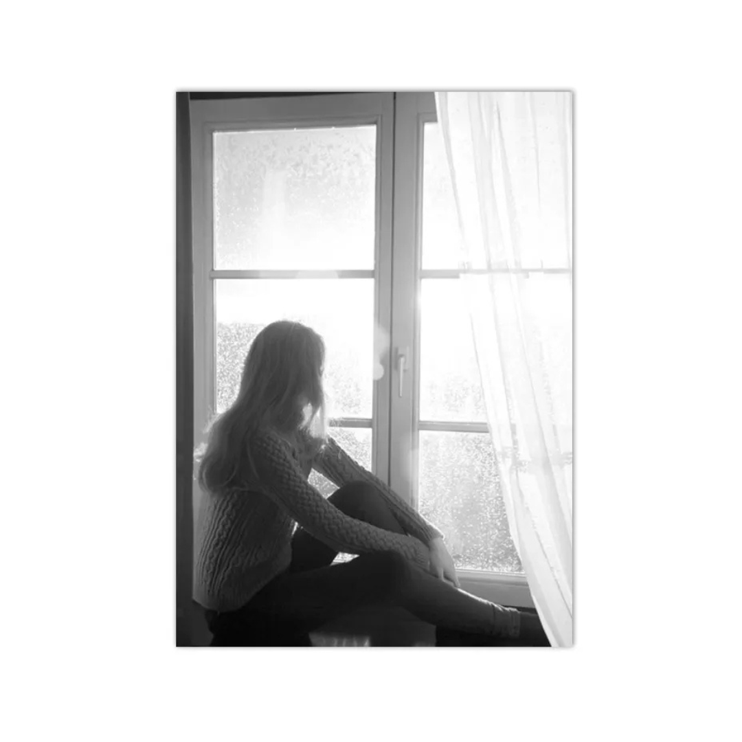 A black and white photographic art print depicting a woman sitting in profile by a window with frosted glass and sheer curtains.