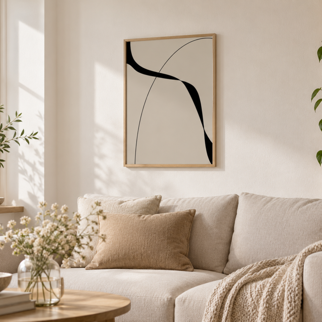 A framed abstract wall art print with black curved lines on a warm off-white background, hanging above a beige sofa in a softly lit living room with pillows, a throw blanket, and a small round table in the foreground.