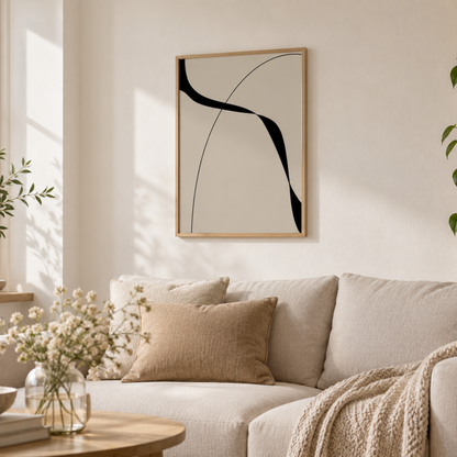 A framed abstract wall art print with black curved lines on a warm off-white background, hanging above a beige sofa in a softly lit living room with pillows, a throw blanket, and a small round table in the foreground.