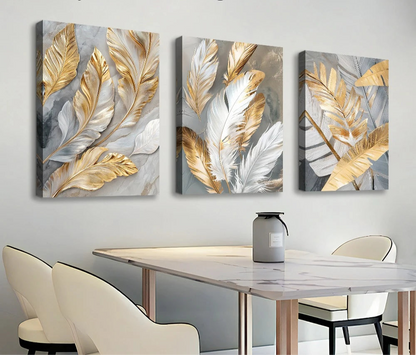 Three abstract wall art pieces with gold and silver leaves on a white wall above a dining table.