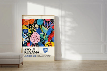 A framed Yayoi Kusama art print leaning against a white paneled wall in a bright room with sunlight and a light gray bench in the foreground.