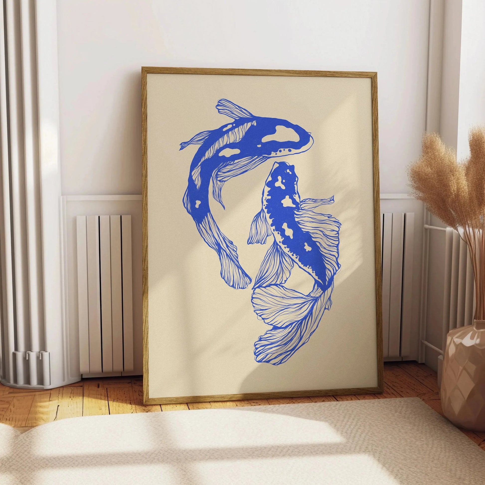 A framed art print featuring two blue koi fish on a cream background, leaning against a white wall in a sunlit room with wood floors and a vase of dried grass.