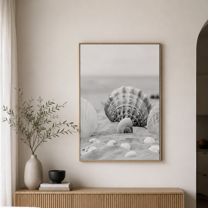 A framed black and white beach print featuring seashells on sand, displayed on a light beige wall above a wooden console table with a vase and small bowl.
