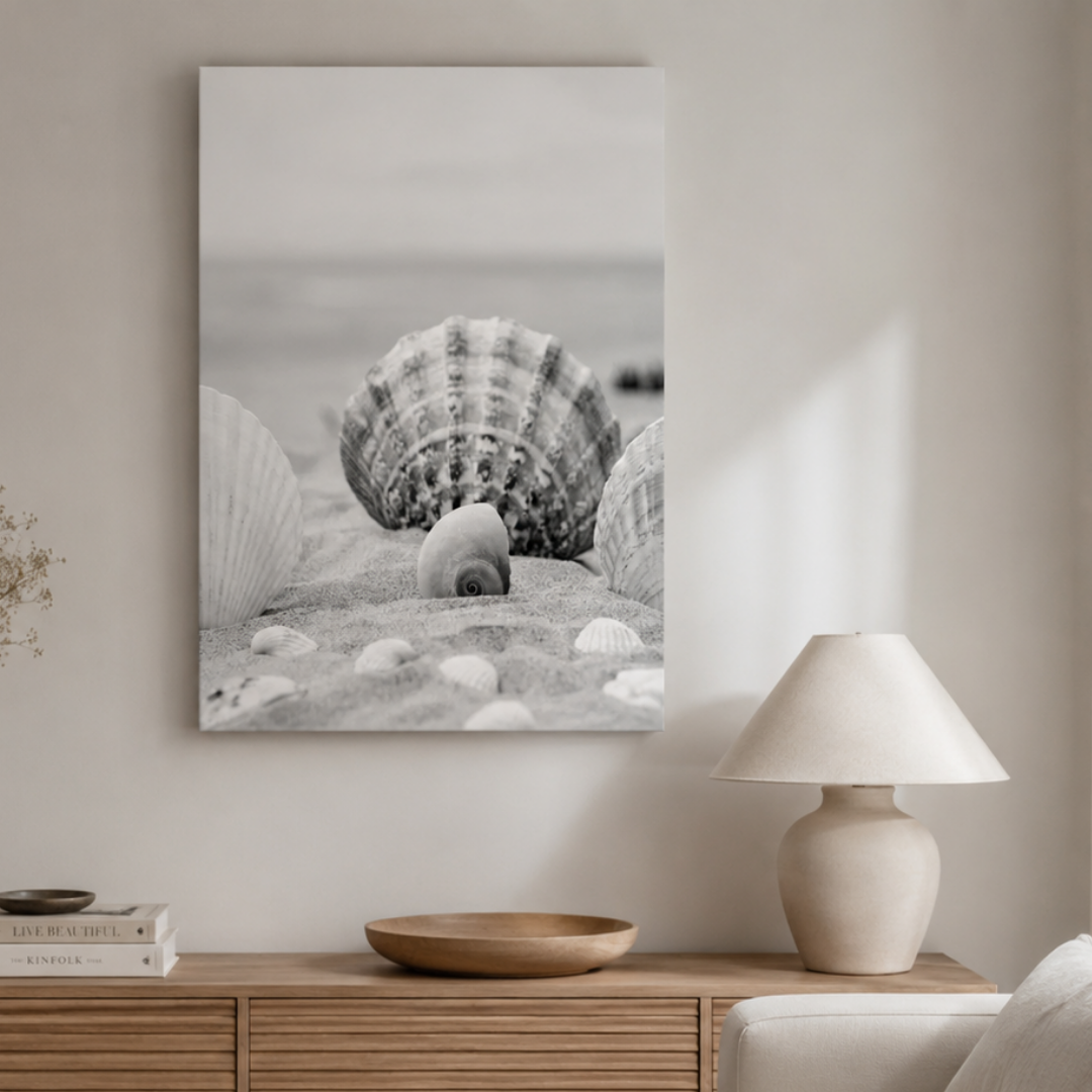 A vertical black and white canvas print of seashells on sand, displayed on a light beige wall above a wooden console table with a lamp and books.